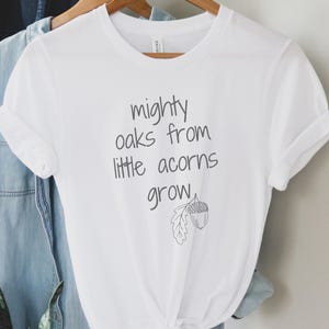 May include: White t-shirt with the text "mighty oaks from little acorns grow" in black script. The shirt features a small acorn and leaf illustration. A light blue denim shirt hangs behind it on a wooden hanger.