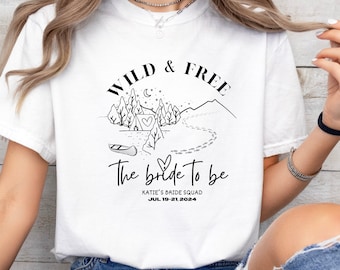 May include: White t-shirt with a black graphic design of a mountain range, a tent, a canoe, and footprints. The text "Wild & Free" is above the graphic and "The bride to be" is below the graphic. The text "Katie's Bride Squad Jul. 19-21, 2024" is below the graphic.