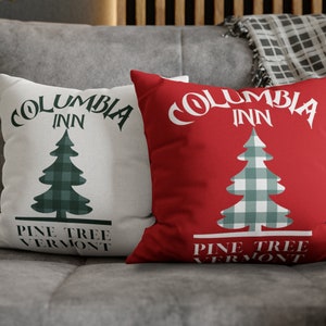 May include: Two decorative throw pillows with the text "Columbia Inn" and a green and white plaid Christmas tree design. The pillows are on a gray couch with a plaid throw blanket.