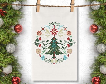 以下が含まれることがあります： 赤い、青い、黄色の花に囲まれた緑色のクリスマスツリーが描かれた白いキッチンクロス。洗濯ばさみで物干しロープにかけられています。