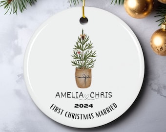 Puede incluir: Adorno navideño redondo blanco con una ilustración de acuarela de un árbol de Navidad en maceta decorado con bayas rojas y adornos dorados. El texto "Amelia ♥ Chris 2024 First Christmas Married" está impreso debajo del árbol.