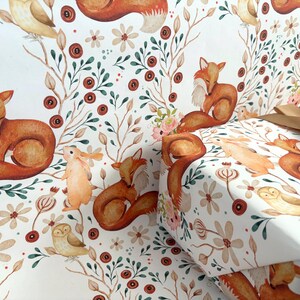 May include: White wrapping paper with a repeating pattern of foxes, owls, and rabbits among flowers and foliage. The gift wrap is tied with a gold ribbon. The overall design has a watercolor effect.