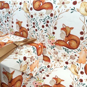 May include: Gift wrap featuring a repeating pattern of foxes, owls, and rabbits among floral and botanical elements. The gift box is wrapped in the same paper and tied with a gold ribbon. The color palette includes shades of brown, orange, and cream.