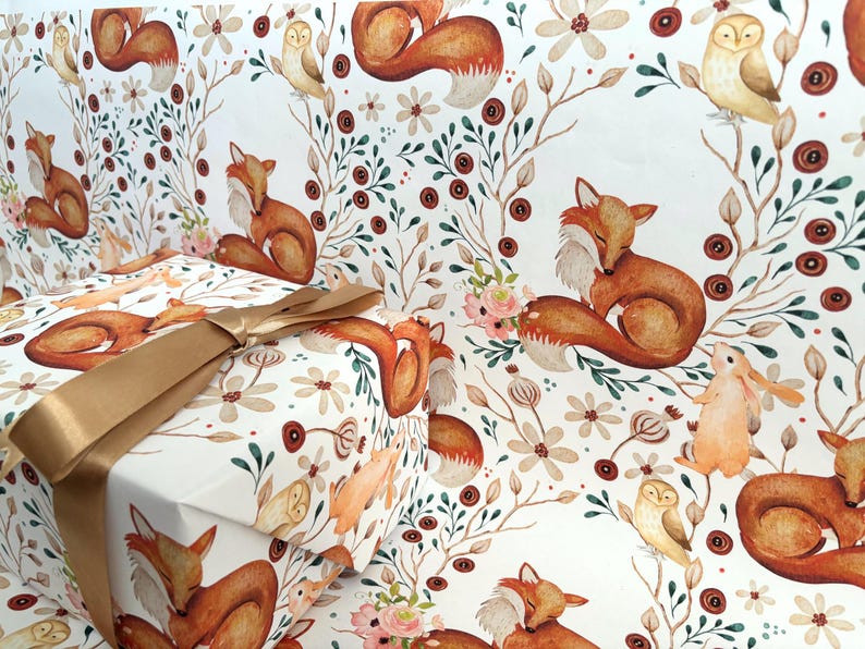 May include: Gift wrap featuring a repeating pattern of foxes, owls, and rabbits among floral and botanical elements. The gift box is wrapped in the same paper and tied with a gold ribbon. The color palette includes shades of brown, orange, and cream.