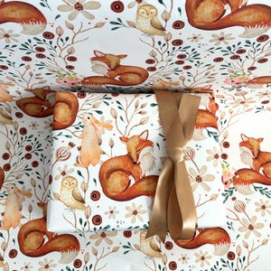 May include: Gift wrapped in white paper with a repeating pattern of foxes, owls, and rabbits in a woodland scene. The gift is tied with a gold ribbon. The wrapping paper features floral and botanical elements.
