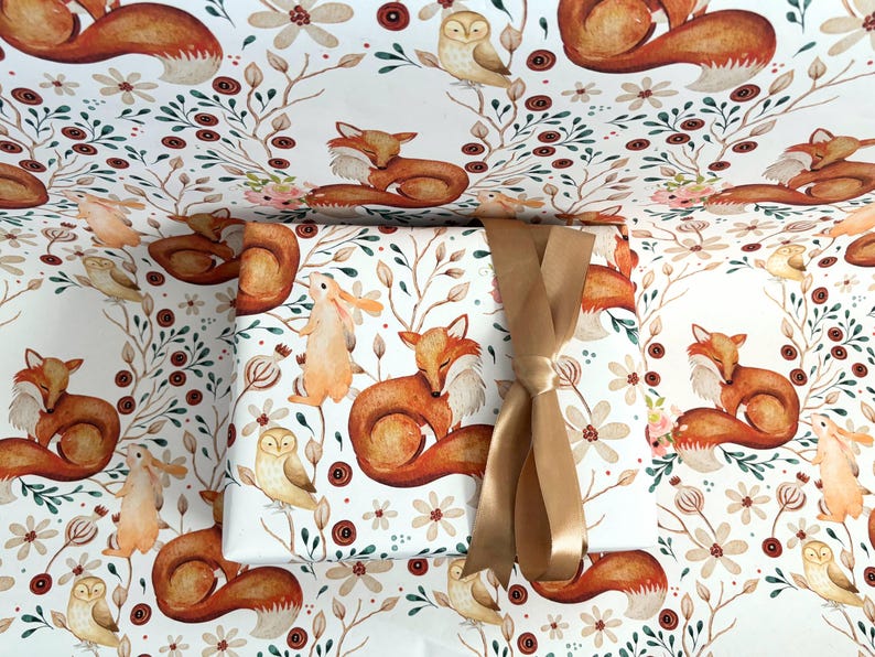 May include: Gift wrapped in white paper with a repeating pattern of foxes, owls, and rabbits in a woodland scene. The gift is tied with a gold ribbon. The wrapping paper features floral and botanical elements.