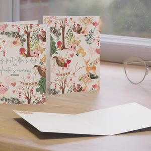 May include: Greeting cards with a whimsical forest design. The cards feature illustrations of woodland creatures, trees, flowers, and hearts. One card reads "happy first mother's day as Hunter's mommy 2026". The cards are open and displayed on a wooden surface.