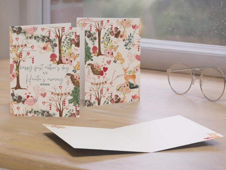 May include: Greeting cards with a whimsical forest design. The cards feature illustrations of woodland creatures, trees, flowers, and hearts. One card reads "happy first mother's day as Hunter's mommy 2026". The cards are open and displayed on a wooden surface.