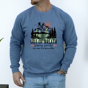 May include: A blue sweatshirt with a graphic of a mountain range, a compass, and the text "OUT OF BREATH hiking society DON'T WORRY I'LL BE THERE IN A MINUTE".