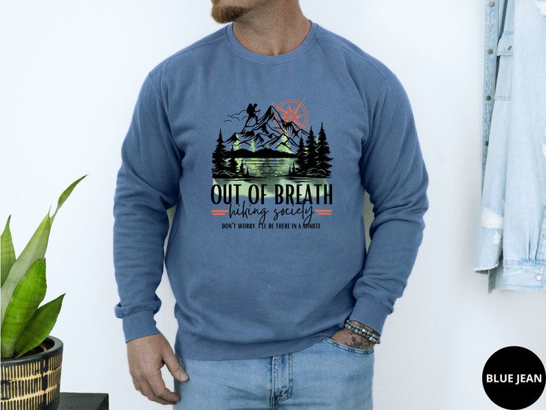 May include: A blue sweatshirt with a graphic of a mountain range, a compass, and the text "OUT OF BREATH hiking society DON'T WORRY I'LL BE THERE IN A MINUTE".