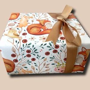 May include: A wrapped gift box featuring a woodland animal print with foxes, owls, and rabbits. The gift is wrapped in white paper with a repeating pattern of forest creatures and floral accents. A gold satin ribbon is tied in a bow.