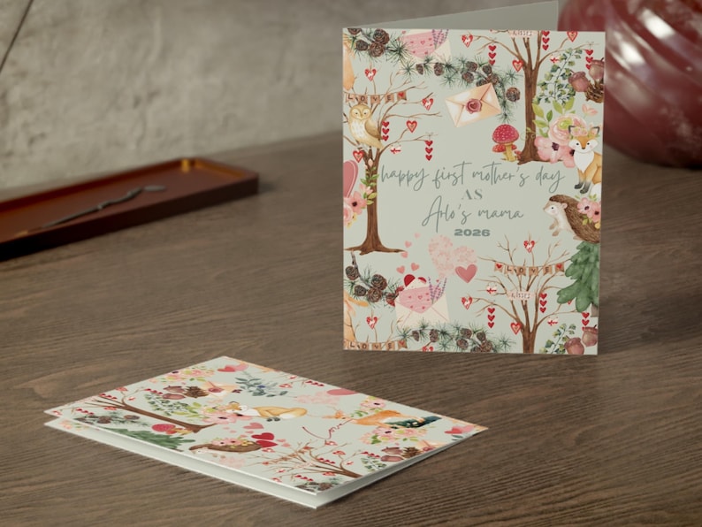 May include: A greeting card with a floral and woodland design, featuring a fox, birds, and hearts. The card reads "Happy First Mother's Day" and "Aria's Mama 2026". A second card with a similar design is visible.