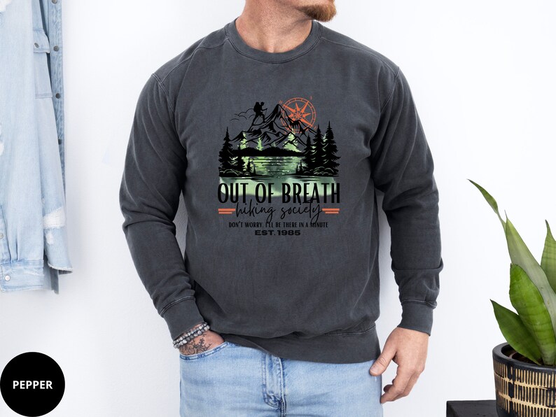 May include: A dark gray sweatshirt with a vintage-style graphic of a mountain scene with a hiker, a compass, and a lake. The text "OUT OF BREATH hiking society DON'T WORRY BE THERE IN A MINUTE EST. 1985" is printed below the graphic.