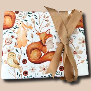 May include: A square, white fabric with a watercolor woodland animal print, featuring a fox, owl, and rabbit. The fabric is tied with a gold ribbon. The design includes floral and botanical elements.