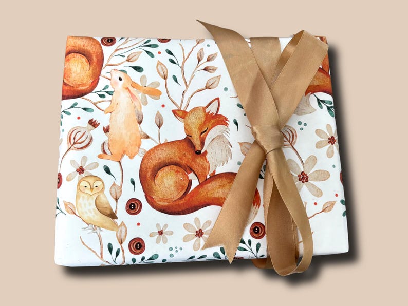 May include: A square, white fabric with a watercolor woodland animal print, featuring a fox, owl, and rabbit. The fabric is tied with a gold ribbon. The design includes floral and botanical elements.