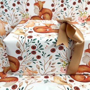 May include: A wrapped gift box featuring a woodland-themed design with foxes, owls, and floral elements. The gift is wrapped in white paper with a repeating pattern and tied with a gold ribbon.