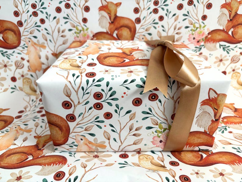 May include: A wrapped gift box featuring a woodland-themed design with foxes, owls, and floral elements. The gift is wrapped in white paper with a repeating pattern and tied with a gold ribbon.