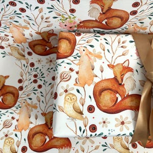 May include: White wrapping paper with a watercolor woodland design featuring foxes, owls, and rabbits. The paper is decorated with floral and botanical elements in shades of brown, orange, and green. A gold ribbon is tied around a wrapped package.