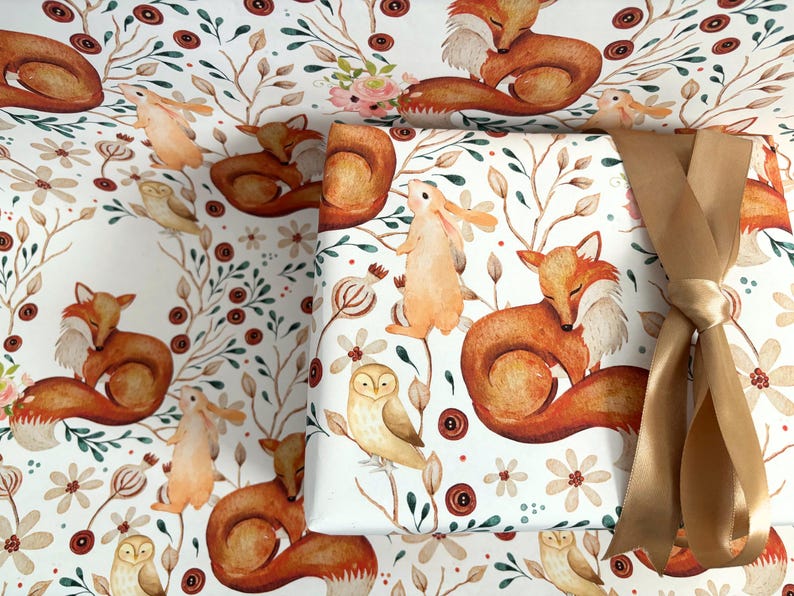 May include: White wrapping paper with a watercolor woodland design featuring foxes, owls, and rabbits. The paper is decorated with floral and botanical elements in shades of brown, orange, and green. A gold ribbon is tied around a wrapped package.