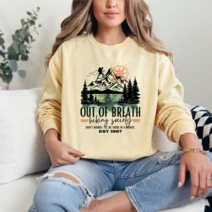 May include: A yellow sweatshirt with a graphic print of a mountain scene and the text "OUT OF BREATH =hiking society= DON'T WORRY. I'LL BE THERE IN A MINUTE EST. 1987"