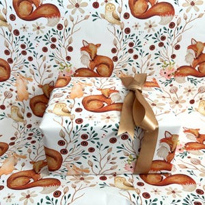 May include: A wrapped gift with a gold ribbon bow, featuring a repeating pattern of watercolor foxes, owls, and floral elements on white wrapping paper. The design includes brown, orange, and green accents.