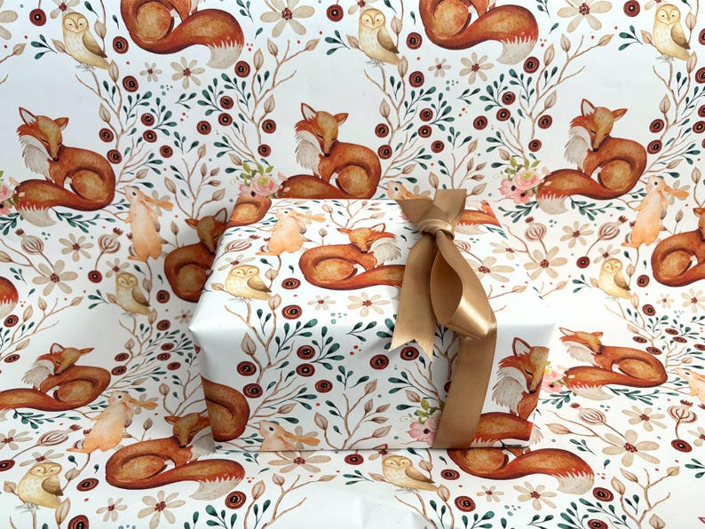 May include: A wrapped gift with a gold ribbon bow, featuring a repeating pattern of watercolor foxes, owls, and floral elements on white wrapping paper. The design includes brown, orange, and green accents.
