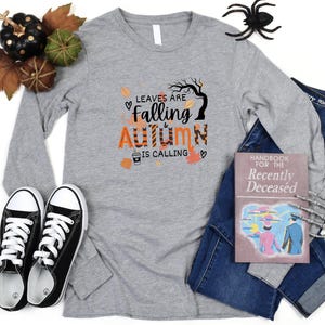 May include: A long-sleeved gray t-shirt with the text "Leaves are Falling & Autumn is Calling" in orange and black lettering. The shirt features a graphic of a tree with leaves falling and a cup of coffee.