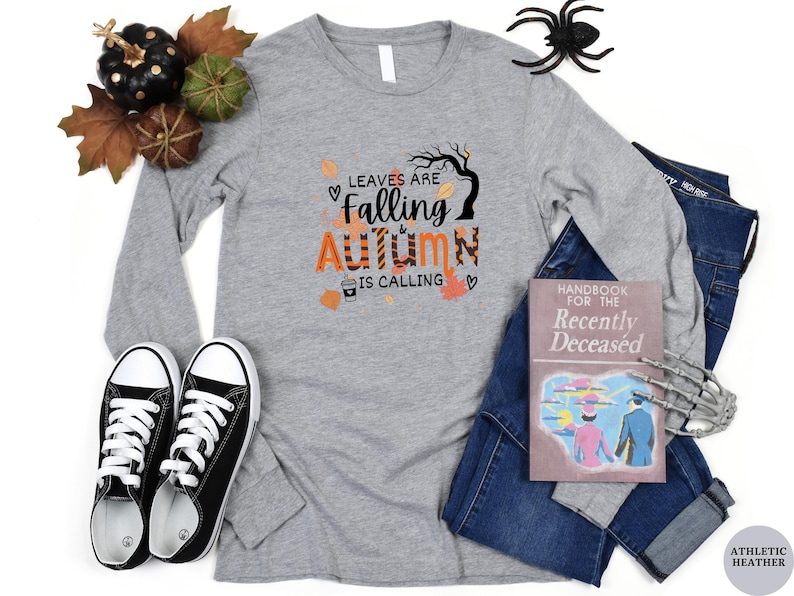 May include: A long-sleeved gray t-shirt with the text "Leaves are Falling & Autumn is Calling" in orange and black lettering. The shirt features a graphic of a tree with leaves falling and a cup of coffee.