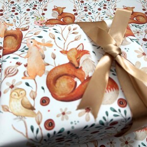 May include: A wrapped gift with a woodland animal print featuring foxes, owls, and rabbits. The gift is tied with a gold satin ribbon. The wrapping paper has a white background with a repeating pattern of forest creatures and floral elements.