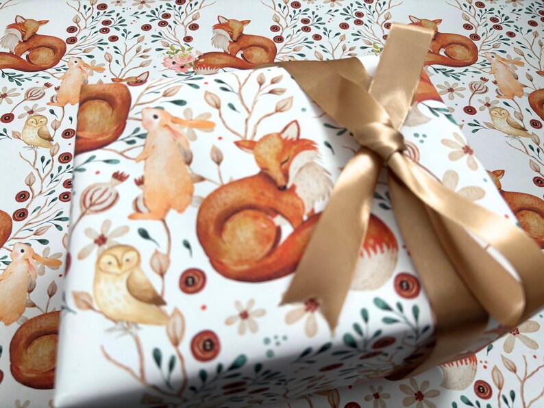 May include: A wrapped gift with a woodland animal print featuring foxes, owls, and rabbits. The gift is tied with a gold satin ribbon. The wrapping paper has a white background with a repeating pattern of forest creatures and floral elements.