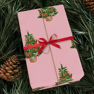 以下が含まれることがあります： 赤いリボンと紐で結ばれた、ピンク色の包装紙。クリスマスツリーが繰り返し描かれており、赤いリボンとオーナメントで飾られています。