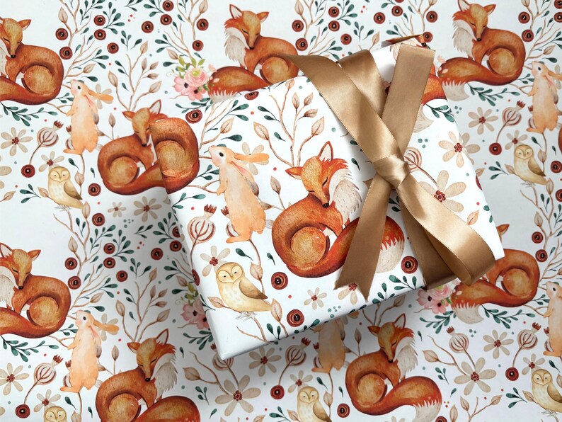 May include: A wrapped gift with a gold ribbon bow sits atop wrapping paper featuring a repeating pattern of watercolor foxes, owls, and rabbits among floral and botanical elements in shades of brown, orange, and green on a white background.