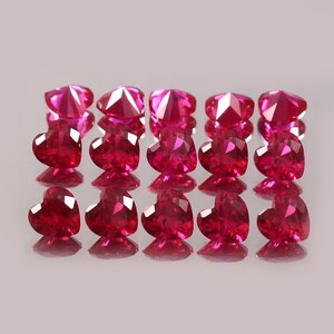 May include: A collection of 15 heart-shaped ruby gemstones, arranged in three rows of five. The rubies are a deep, rich red colour and have a high level of brilliance.