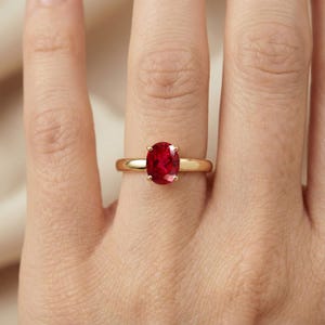 Könnte beinhalten: Ein Goldring mit einem einzelnen oval geschliffenen roten Edelstein in einer Vier-Krappen-Fassung. Die Ringschiene ist glatt und poliert und zeigt ein klassisches Design, das die leuchtende Farbe und Klarheit des Steins am Finger betont.