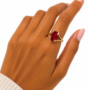 Puede incluir: Un anillo de oro con una gran piedra preciosa de color rojo intenso y corte ovalado, montada en un engaste sencillo. La banda del anillo es fina y pulida, mostrando un diseño clásico que resalta el color vibrante y la claridad de la piedra.