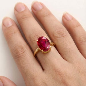 Peut inclure: Une bague en or avec une grande pierre précieuse rouge de taille ovale, fixée par quatre griffes. L'anneau est en or jaune poli, et le sertissage maintient fermement la pierre rouge à facettes, révélant des tons cramoisis et des reflets.