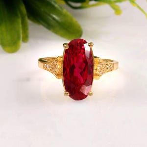 Pode incluir: Um anel de ouro com uma grande pedra preciosa de corte oval e cor vermelho-profundo, presa por quatro garras. A banda é feita de ouro amarelo e inclui pequenas pedras transparentes incrustadas em ambos os lados da pedra central.