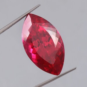 May include: A faceted, marquise-cut gemstone held by tweezers. The gem is a vibrant, deep red color with multiple facets that reflect light. The shape is elongated and pointed at both ends, showcasing its polished surface.