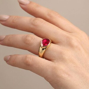 Peut inclure: Une bague en or ornée d'une pierre précieuse rouge vif en forme de poire, montée sur un anneau lisse et poli. Le sertissage est de type clos, fixant la pierre en forme de goutte au centre de l'anneau en métal doré épais et arrondi.