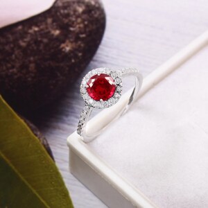 Könnte beinhalten: Ein silberfarbener Ring mit einem runden, leuchtend roten Mittelstein, der von einem Kranz aus kleinen, klaren, funkelnden Steinen umgeben ist. Die Ringschiene ist schmal und ebenfalls mit einer Reihe kleiner, klarer Steine besetzt.