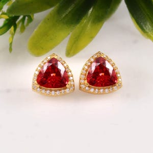 May include: A pair of stud earrings featuring triangular-cut red gemstones set in gold metal. Each central red stone is surrounded by a halo of small, clear white accent stones, creating a bright and reflective appearance on the gold settings.