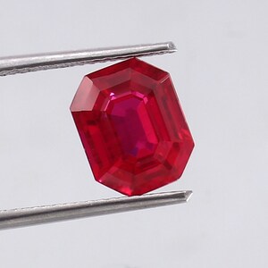 Puede incluir: Una gema roja vibrante con un corte esmeralda rectangular. La gema está sujeta por unas pinzas.