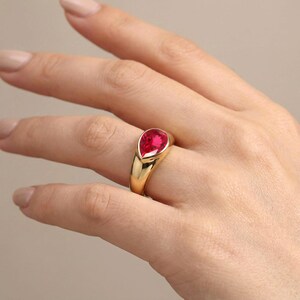 Può includere: Un anello in oro con una singola pietra preziosa rosso vivo a forma di pera, incastonata in una fascia liscia e lucida. La pietra a goccia è fissata in una montatura a castone che si integra perfettamente nella fascia d'oro spessa e arrotondata.