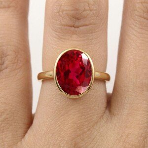 May include: A gold ring featuring a large, oval-cut, vibrant red gemstone set in a smooth bezel setting. The band is polished yellow gold, providing a clean and simple frame for the faceted red stone that catches the light.