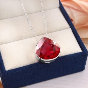 Puede incluir: Un collar de plata con un gran colgante de piedra preciosa roja facetada en forma de lágrima. El colgante está engastado en un bisel de plata y cuelga de una delicada cadena de plata. El collar está en una caja de joyería azul.