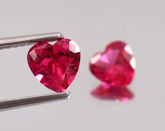 May include: Two heart-shaped, faceted, red gemstones. One is held by silver tweezers, the other is slightly out of focus in the background. The gems have a brilliant, reflective surface, suggesting they are cut from a precious stone.