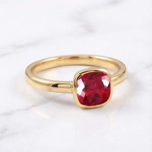 Op de afbeelding: Een gouden ring met een enkele, kussengeslepen, dieprode edelsteen gezet in een gladde, gepolijste metalen zetting. De ringband is slank en afgerond en heeft een hoogglans afwerking die het licht reflecteert op het oppervlak van de rode steen.