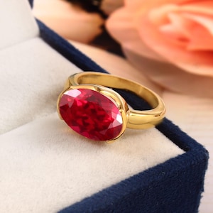 Op de afbeelding: Een gouden ring met een grote, ovaal geslepen, helderrode edelsteen. De edelsteen is gezet in een gepolijste gouden band. De ring wordt gepresenteerd in een donkerblauwe fluwelen sieradendoos, met een zachte, vage achtergrond.