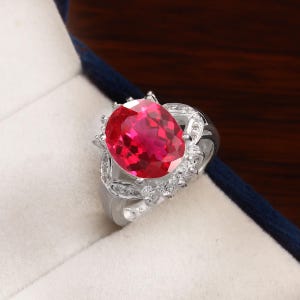 May include: A silver-toned ring featuring a large, oval-cut, vibrant red gemstone as the centerpiece. The setting is adorned with a cluster of small, clear, sparkling stones surrounding the central red gem and along the band's decorative shoulders.