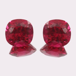 May include: Two cushion-cut, faceted, deep red gemstones. Each gem has a square shape with rounded corners, reflecting light. The gems sit on a reflective surface, enhancing their brilliance. Ideal for jewelry making or collecting.